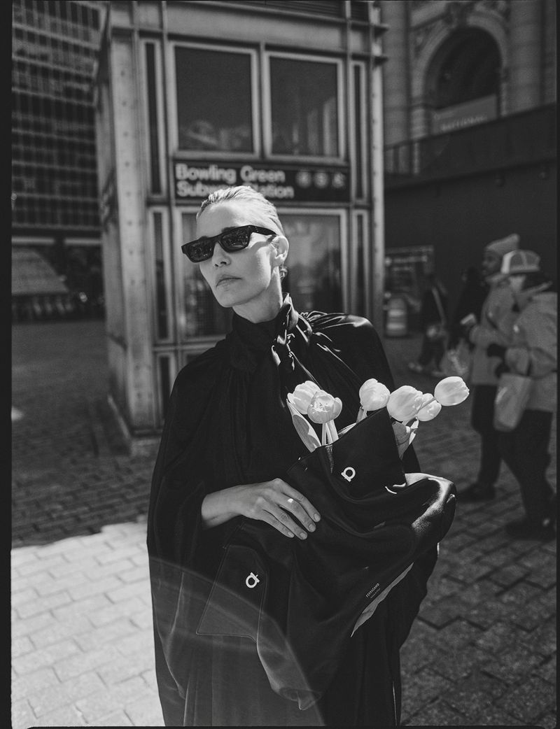 Black and white medium shot of woman in dark clothing carrying tulips in NYC, standing near subway entrance on city sidewalk.