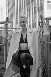 Leslie Bibb smiling while crouching outdoors wearing a trench coat and dark leggings against a high-rise building background.
