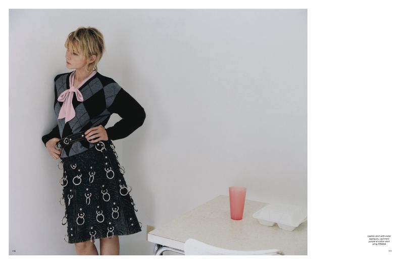 A young adult woman in a fashionable argyle sweater and skirt with silver rings stands against a white wall with a table nearby.