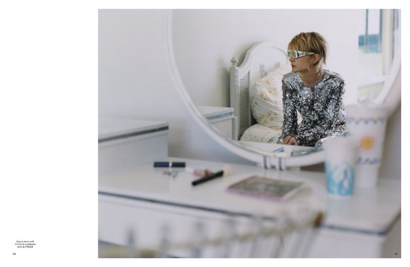 Woman with blonde hair wearing sunglasses and sequin dress is looking at mirror in bedroom with white vanity and floral bedsheets.