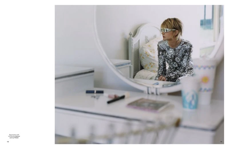 Woman with blonde hair wearing sunglasses and sequin dress is looking at mirror in bedroom with white vanity and floral bedsheets.