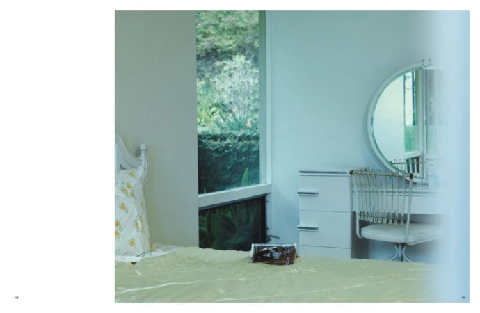 Bedroom interior with a bed, white dresser, round mirror, and chair in a minimalist style. A window offers a view of greenery outside.
