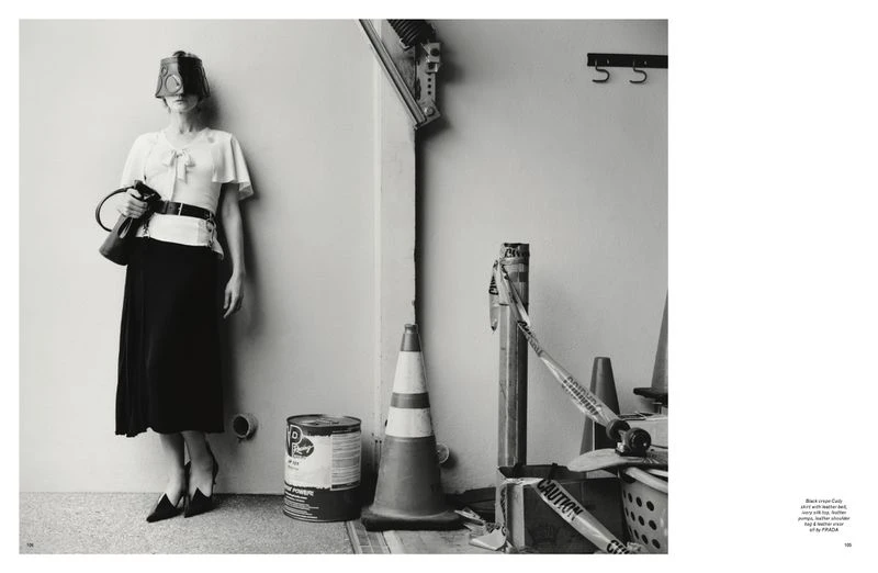 A woman in a surreal black-and-white fashion photograph with a bizarre head covering, posing near construction site objects.