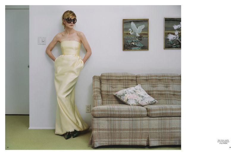 Woman in yellow strapless gown stands in front of couch and art in a minimalist indoor setting, wearing dark sunglasses and posing. 