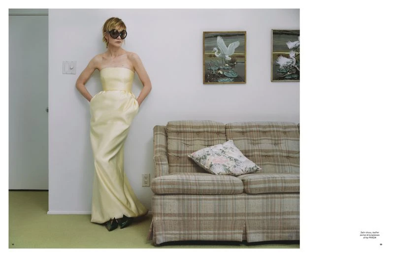 Woman in yellow strapless gown stands in front of couch and art in a minimalist indoor setting, wearing dark sunglasses and posing. 