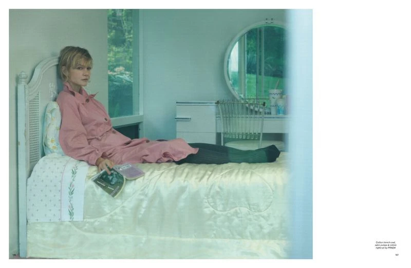 Woman in pink loungewear relaxing on a bed in a brightly lit bedroom with a window and furniture, holding reading material.