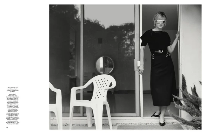 Indoor shot of an adult female model with visor sunglasses, posing in a black dress, entering a building with white plastic chairs.