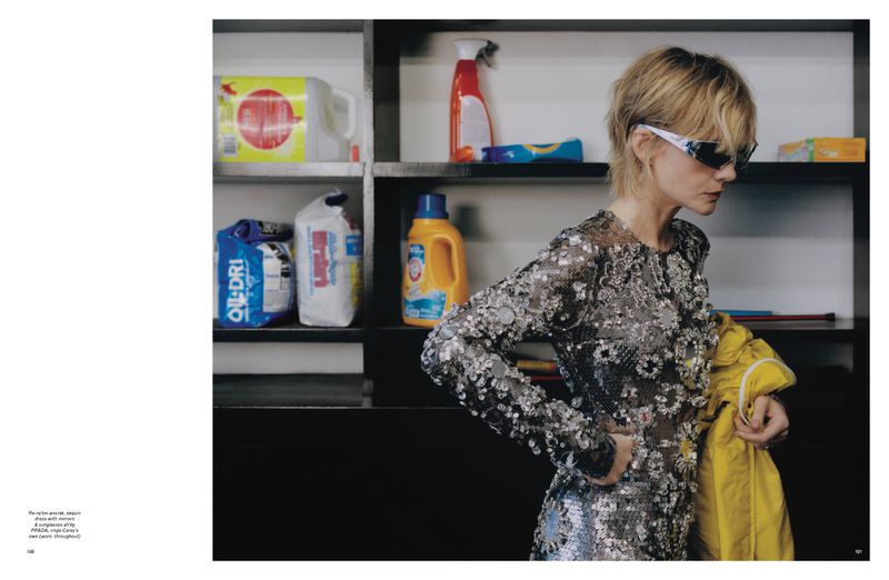 Woman in shimmering dress holds yellow jacket near shelves of cleaning supplies in indoor setting with soft, diffused lighting.