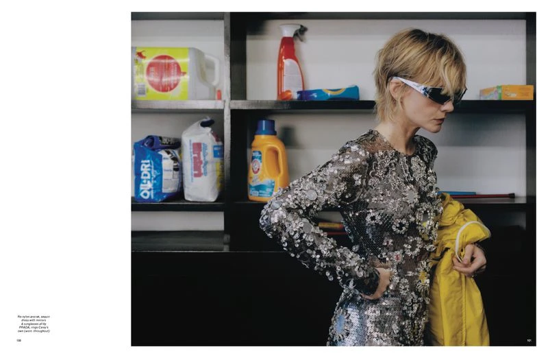 Woman in shimmering dress holds yellow jacket near shelves of cleaning supplies in indoor setting with soft, diffused lighting.