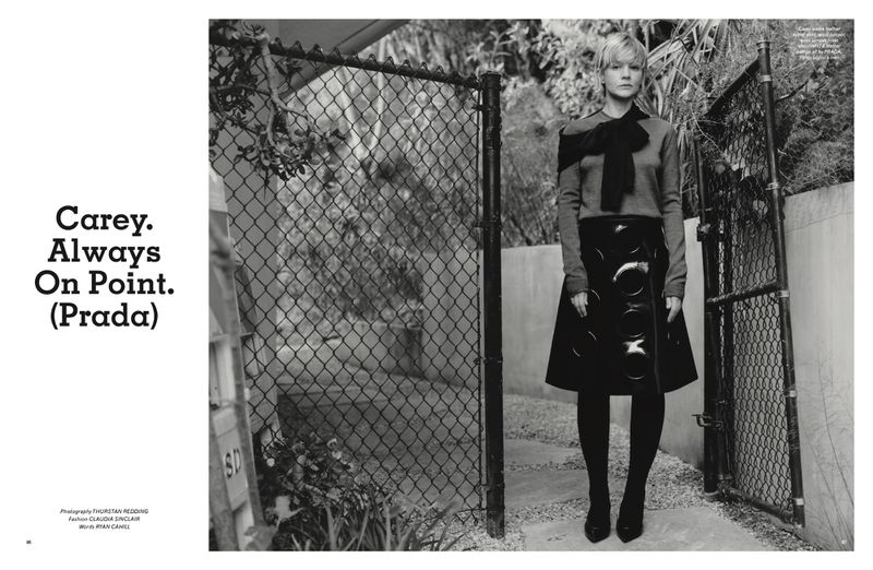 A black and white fashion editorial shot of a woman standing outdoors in a sweater and skirt with design details, next to a fence.