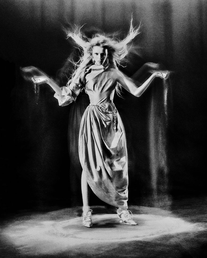 A woman in a draped garment stands in a spotlight, sand cascading from her hands in a black and white studio photograph.