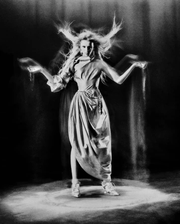 A woman in a draped garment stands in a spotlight, sand cascading from her hands in a black and white studio photograph.