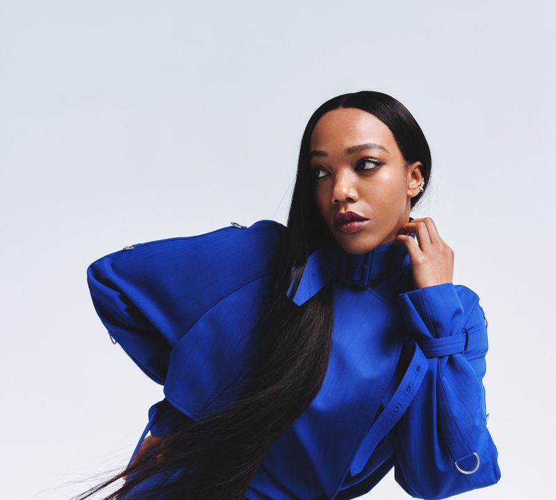 Portrait of an adult woman in a striking blue jacket with long black hair looking off-camera against a neutral studio backdrop.