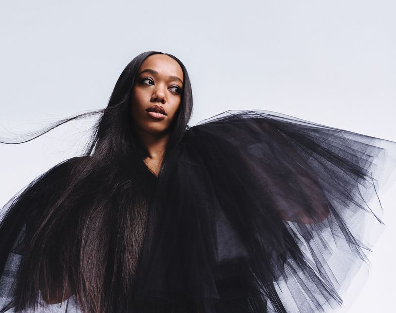 Portrait of a stylish Black woman in black tulle dress. The woman looks off-camera to the right, set against a clean white backdrop.