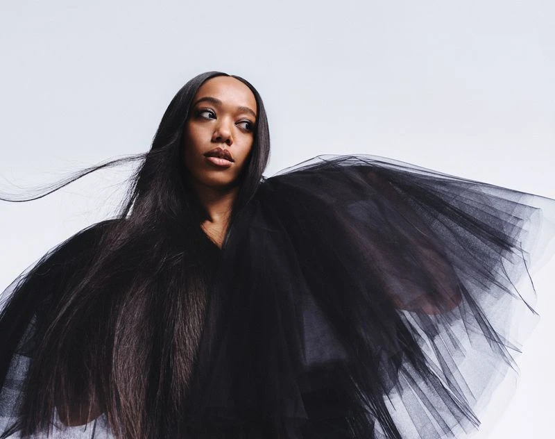 Portrait of a stylish Black woman in black tulle dress. The woman looks off-camera to the right, set against a clean white backdrop.