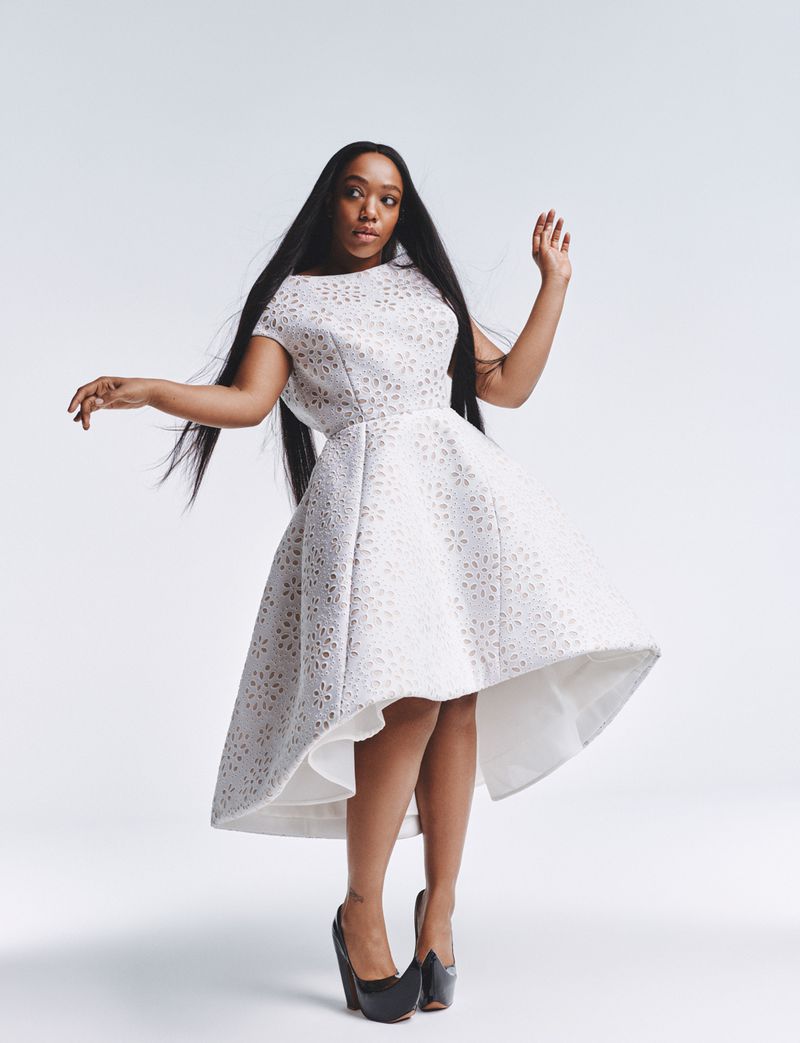 Full-shot portrait of an adult woman in an elegant white dress and black heels, against a bright white studio backdrop, with dark hair.