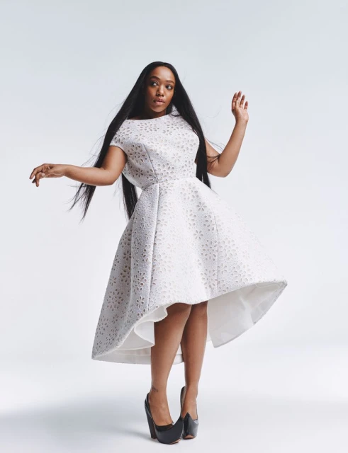 Full-shot portrait of an adult woman in an elegant white dress and black heels, against a bright white studio backdrop, with dark hair.