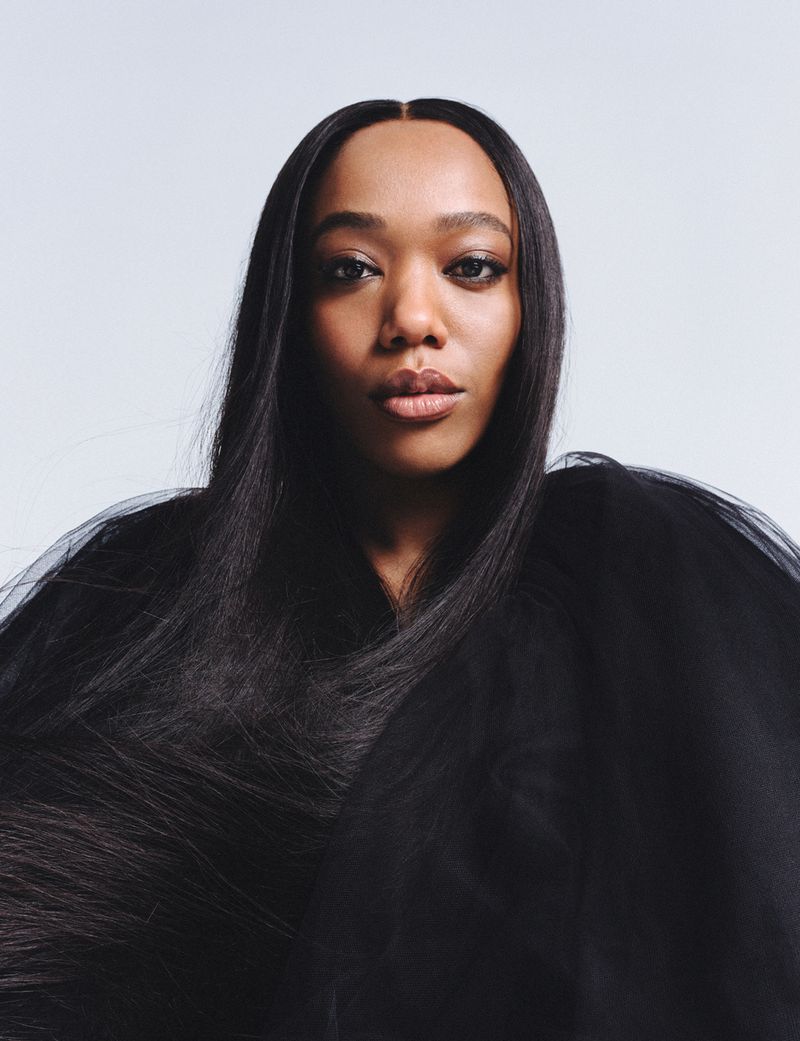 Close-up portrait of a Black adult female with long, straight black hair and a dark translucent garment against a light backdrop.