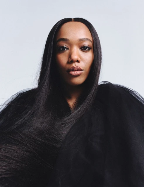 Close-up portrait of a Black adult female with long, straight black hair and a dark translucent garment against a light backdrop.