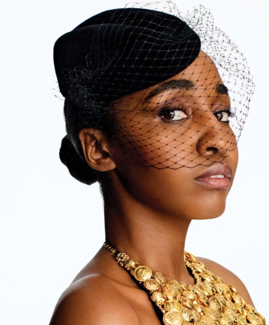Close-up portrait of an African-American woman wearing a black hat with netting and a gold coin necklace on a white background.