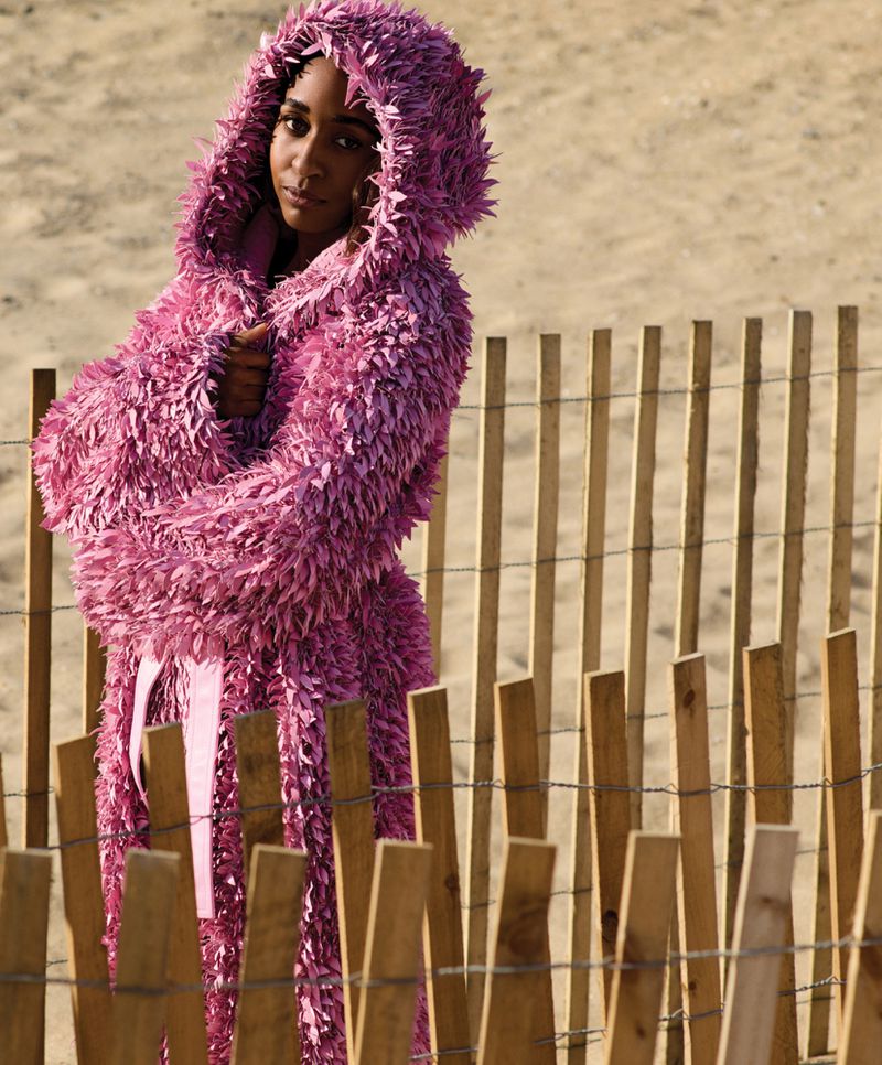 A young adult woman wearing a large, pink coat with a hood stands in front of a weathered wood fence and sandy beach background.
