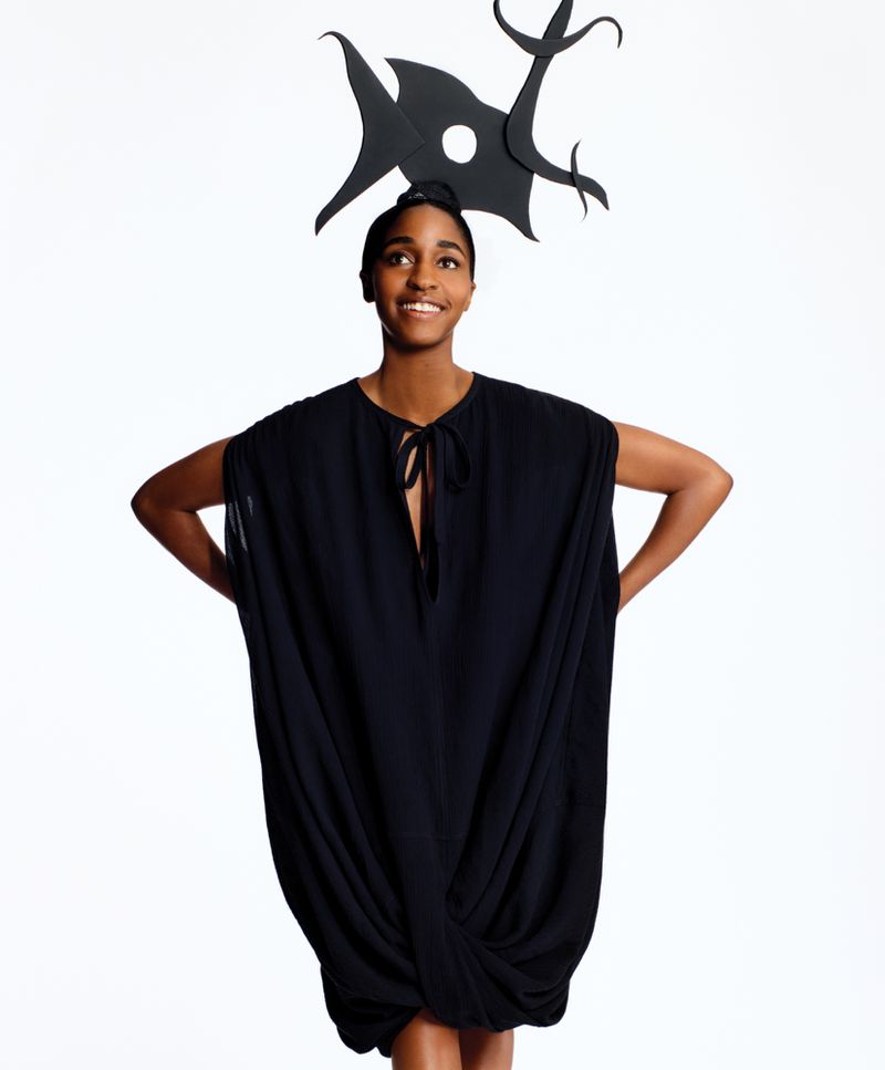A single smiling Black woman poses in a photo studio wearing a black dress and a large black paper headpiece with abstract shapes.