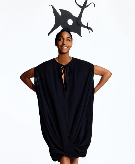 A single smiling Black woman poses in a photo studio wearing a black dress and a large black paper headpiece with abstract shapes.