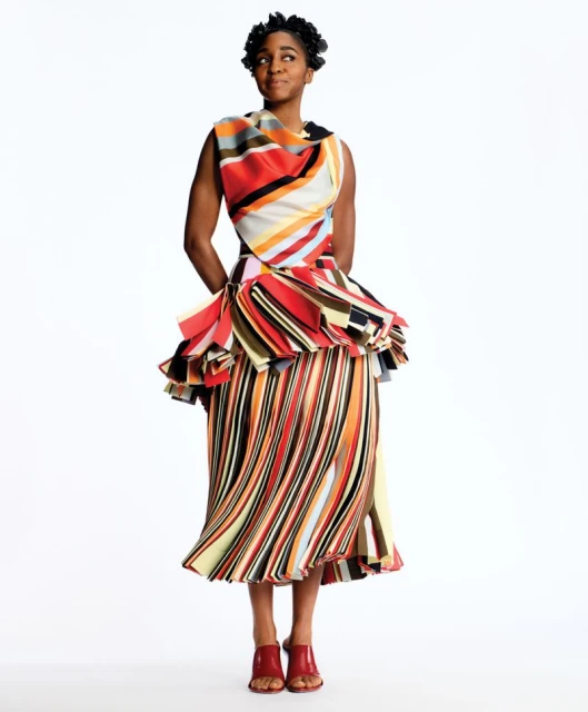 Elegant Black woman posing in a studio wearing a vibrant, striped dress and red shoes against a white background in a full shot.