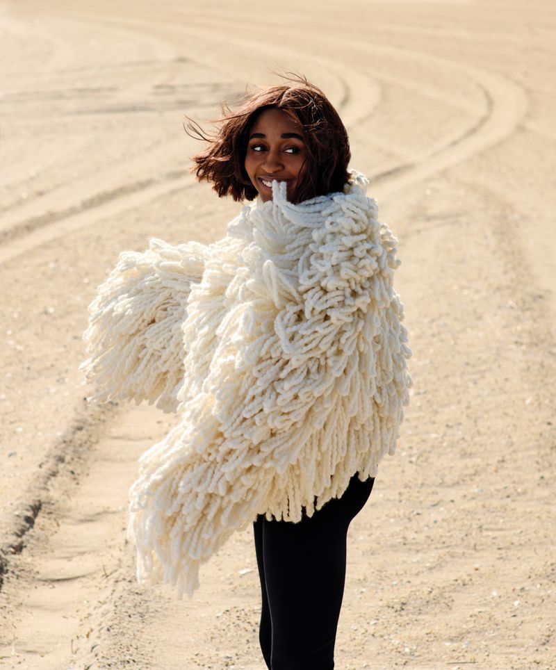 Woman in oversized, textured white coat poses on sandy terrain, smiling at camera. Fashion shot with natural light, neutral tones.