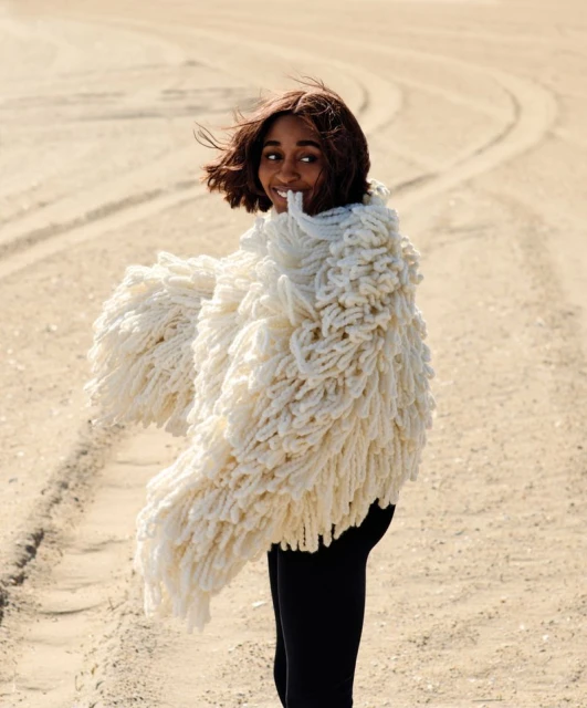 Woman in oversized, textured white coat poses on sandy terrain, smiling at camera. Fashion shot with natural light, neutral tones.