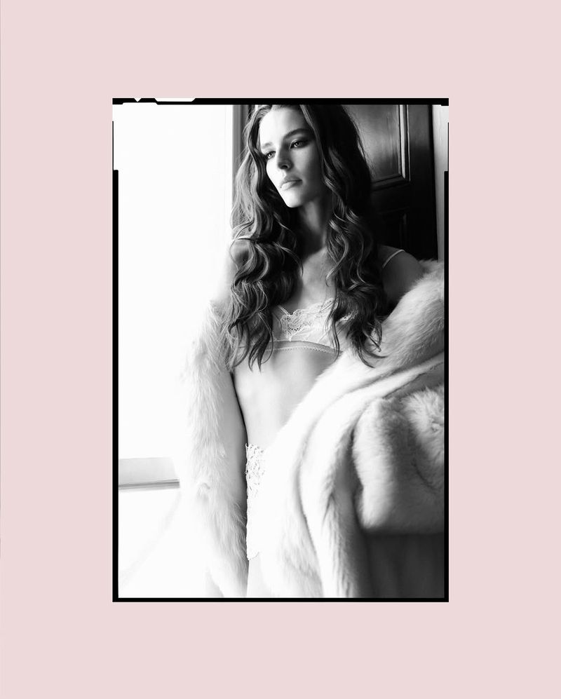 Black and white portrait of a woman with long, wavy hair, wearing lingerie and a fur coat, posing indoors near a window.