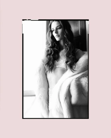 Black and white portrait of a woman with long, wavy hair, wearing lingerie and a fur coat, posing indoors near a window.