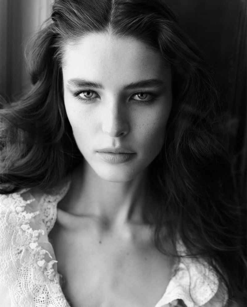 Monochrome portrait of a young fair-skinned woman with long wavy hair, wearing a lace top, gazing directly at the camera indoors.