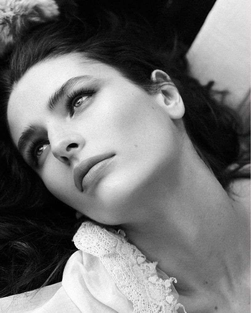 Monochrome portrait of a woman with dark hair looking upwards, wearing a lace-trimmed top, with soft lighting and shallow depth of field.