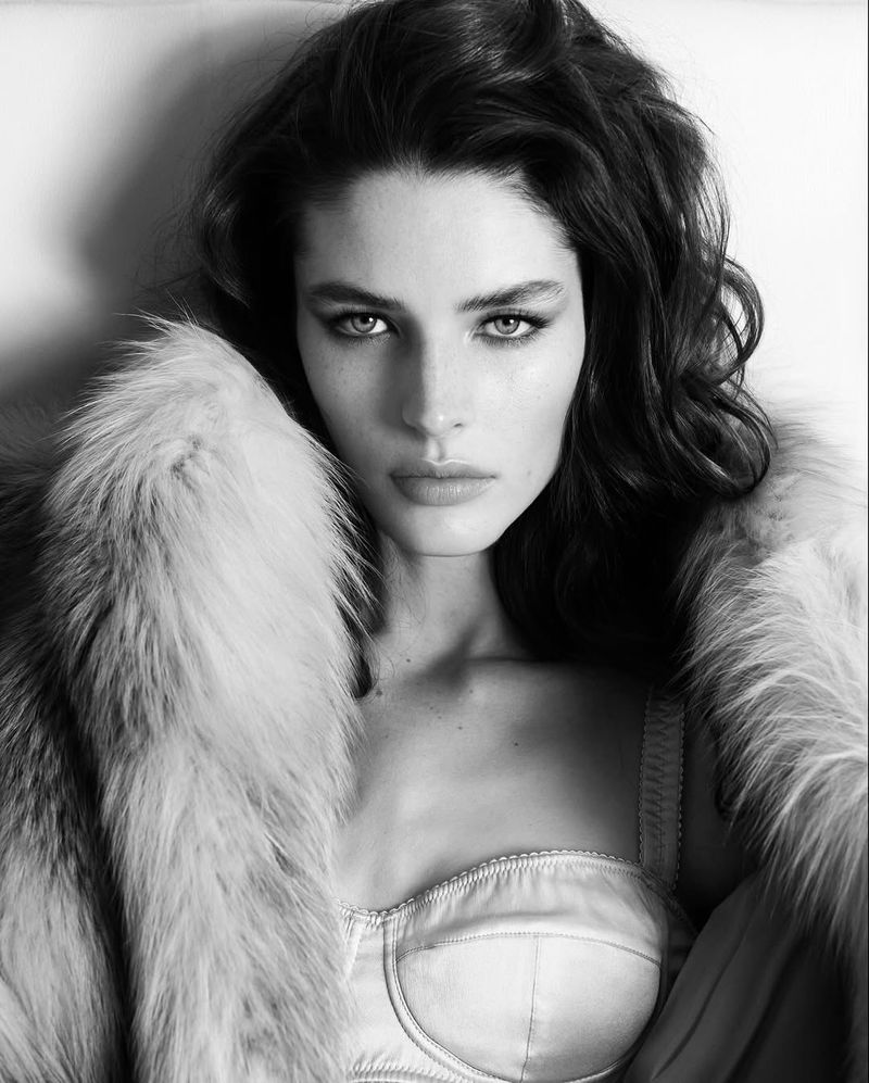 Black and white portrait of a woman with dark wavy hair and pale skin wearing a light bustier top and fur coat, looking at camera.