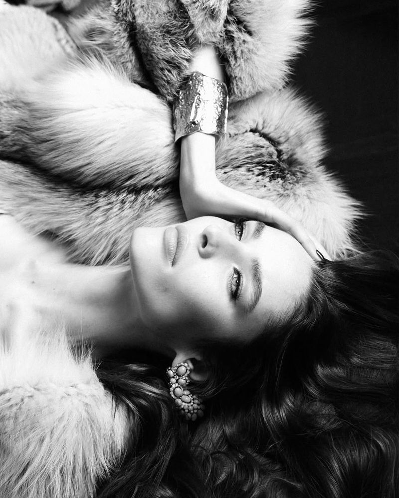 Black and white portrait of a woman lying down wearing a fur coat, statement earrings, and a silver bracelet. Hairstyle is wavy.