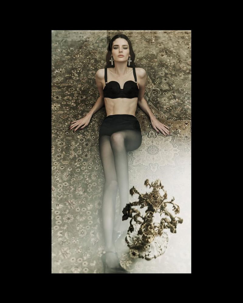 Adult female model in lingerie set and sheer stockings lying on an ornate carpet in a studio setting, accessorized with large earrings.