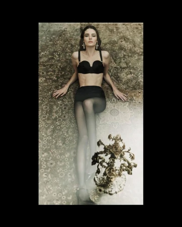 Adult female model in lingerie set and sheer stockings lying on an ornate carpet in a studio setting, accessorized with large earrings.
