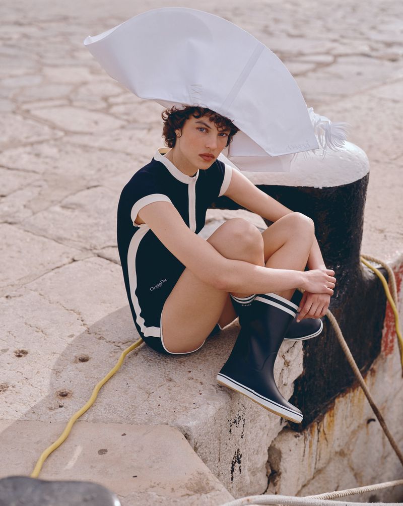 A woman in a swimsuit and rain boots sits on a pier, wearing a large white hat in an outdoor fashion editorial shot.