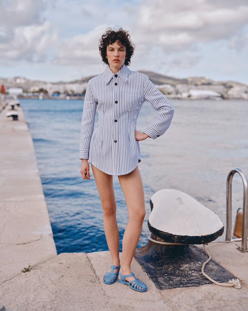 Young adult woman in striped shirt and sandals stands on a concrete pier with ocean and buildings in the background on a bright day.
