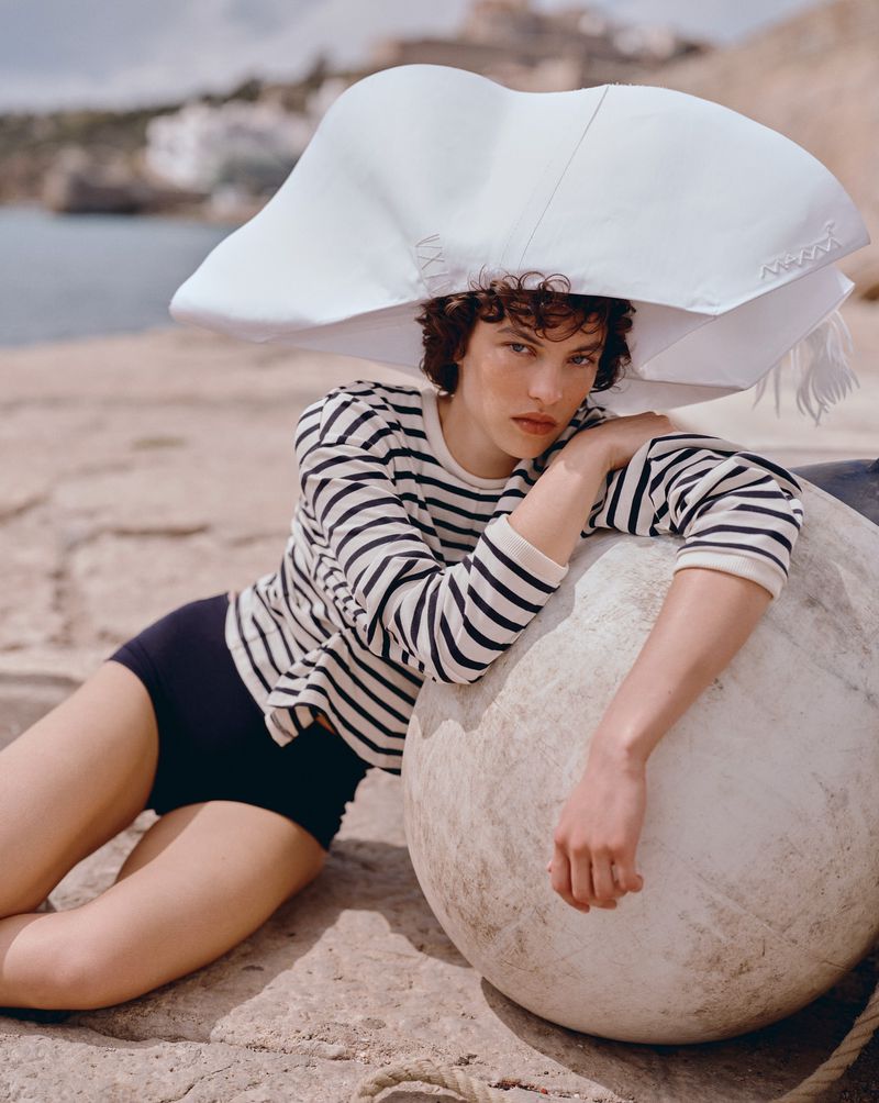 Adult wearing striped shirt, white hat, and black shorts poses with buoy on a stone surface near the ocean and coastal buildings.