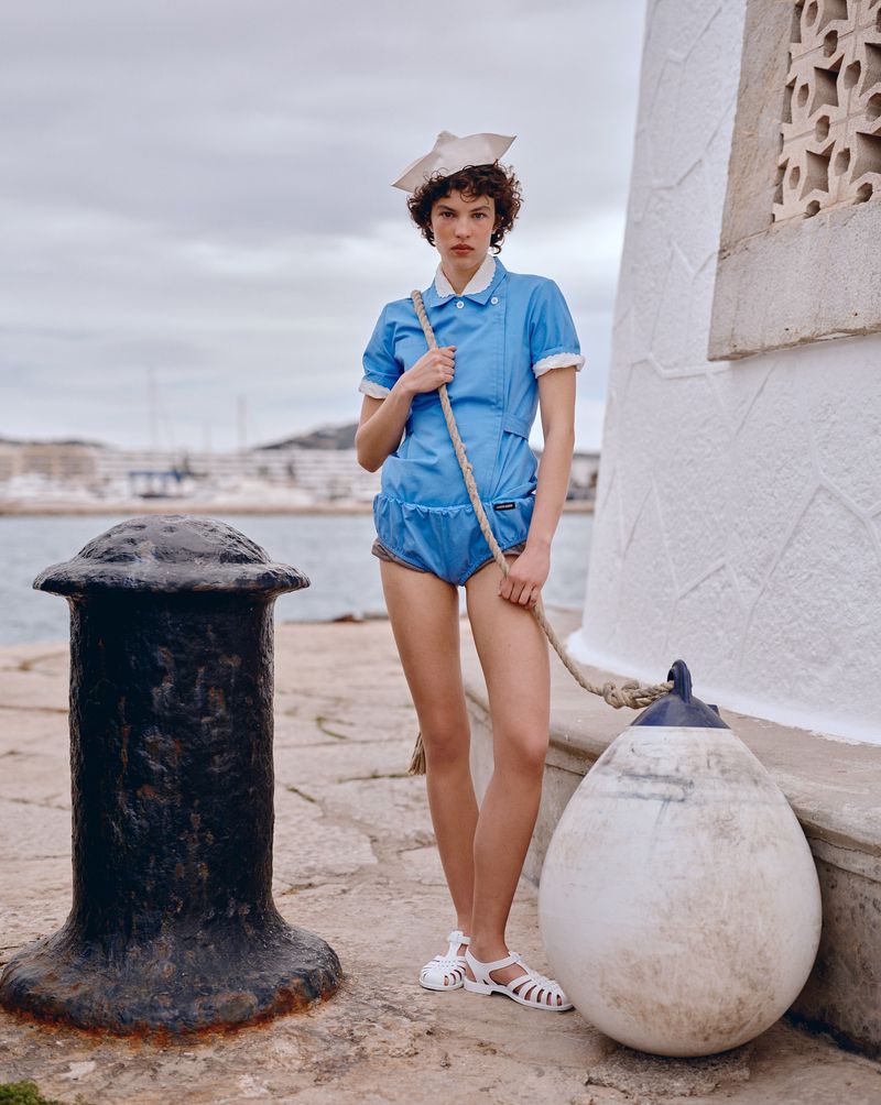 Fashionable woman in blue romper and sailor hat poses on a pier with a nautical rope and mooring buoy against a harbour backdrop.