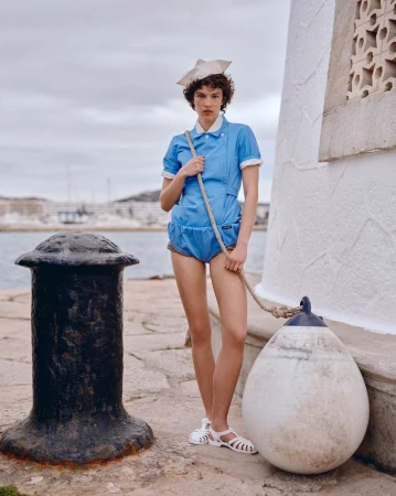 Fashionable woman in blue romper and sailor hat poses on a pier with a nautical rope and mooring buoy against a harbour backdrop.