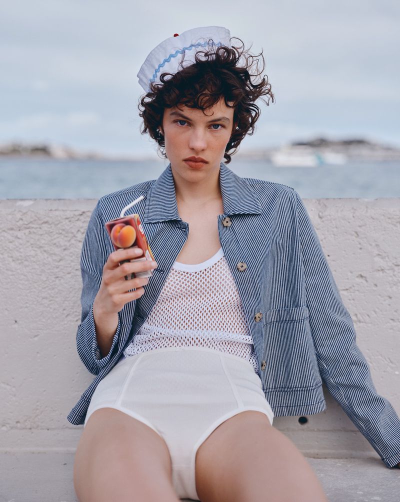 Female model poses outdoors wearing retro white swimsuit bottoms and blue striped jacket, holding fruit juice box with drinking straw.