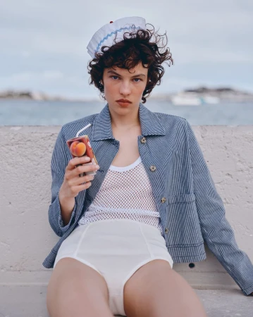 Female model poses outdoors wearing retro white swimsuit bottoms and blue striped jacket, holding fruit juice box with drinking straw.