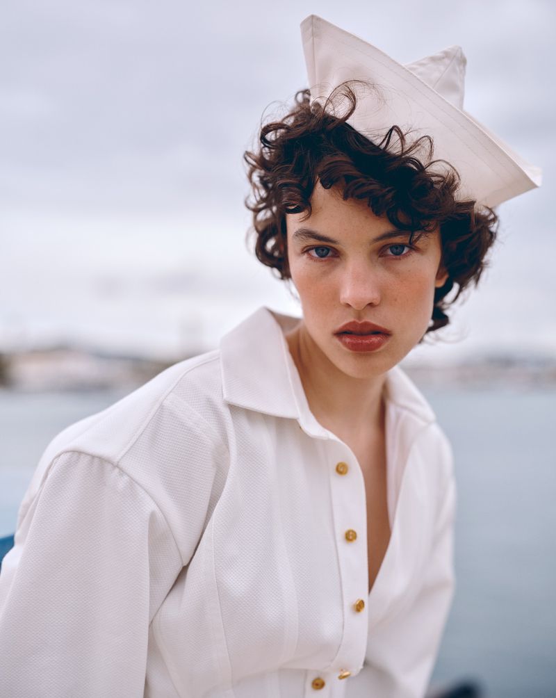 Portrait of a young woman with curly hair wearing a white sailor hat and white collared shirt with gold buttons, posing outdoors near water.