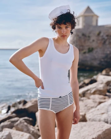 Young woman in a sailor cap and striped underwear poses outdoors with rocks and ocean views, enjoying a sunlit moment.