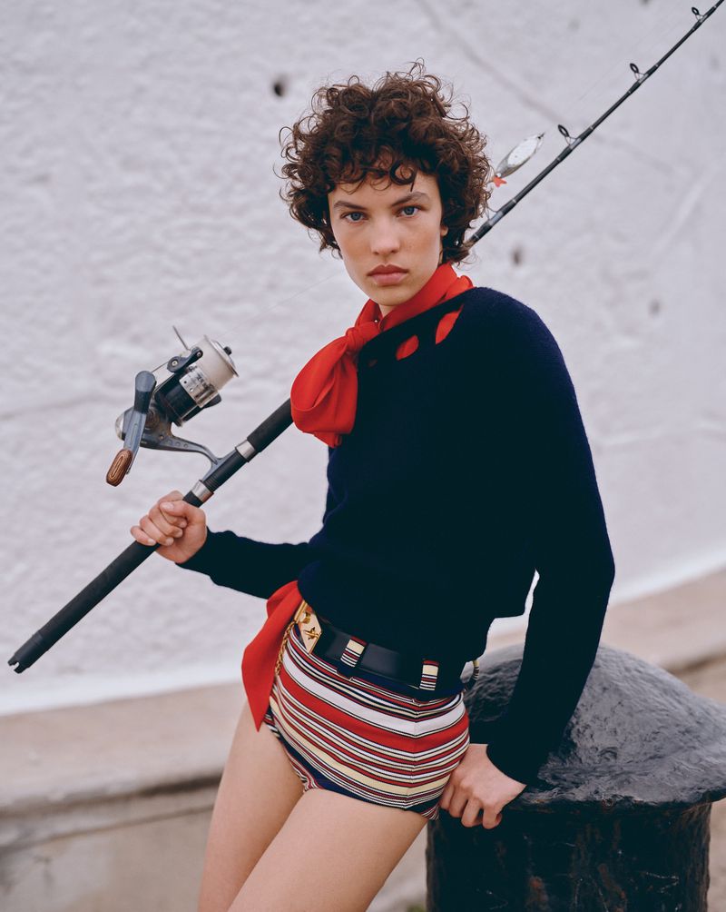 Young woman in striped shorts and dark top poses with fishing rod and reel against a white wall backdrop, captured in natural light.