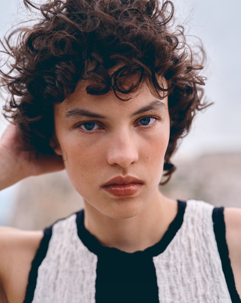 Close-up studio shot of a woman with curly brown hair and blue eyes, wearing a black and white top, against a neutral background.