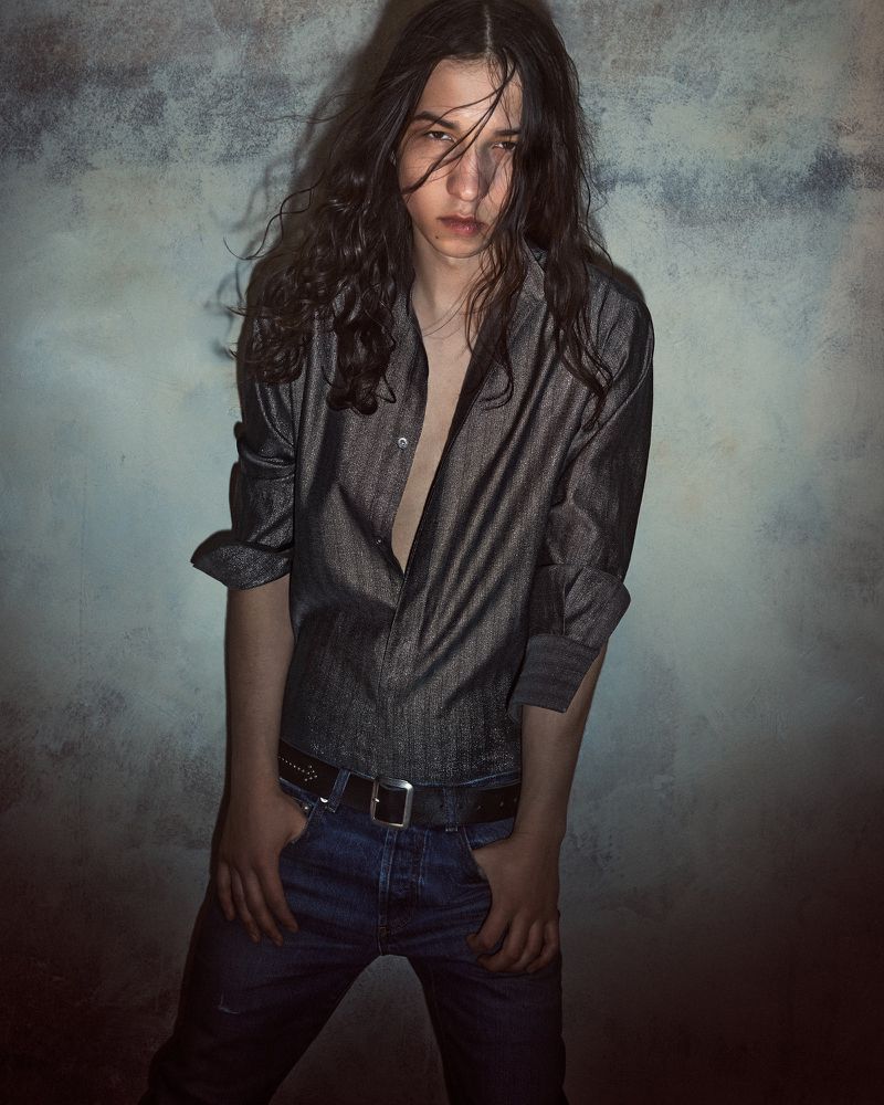 Portrait of a young adult male with long wavy hair wearing an unbuttoned dark gray shirt and jeans, posing against a textured wall.